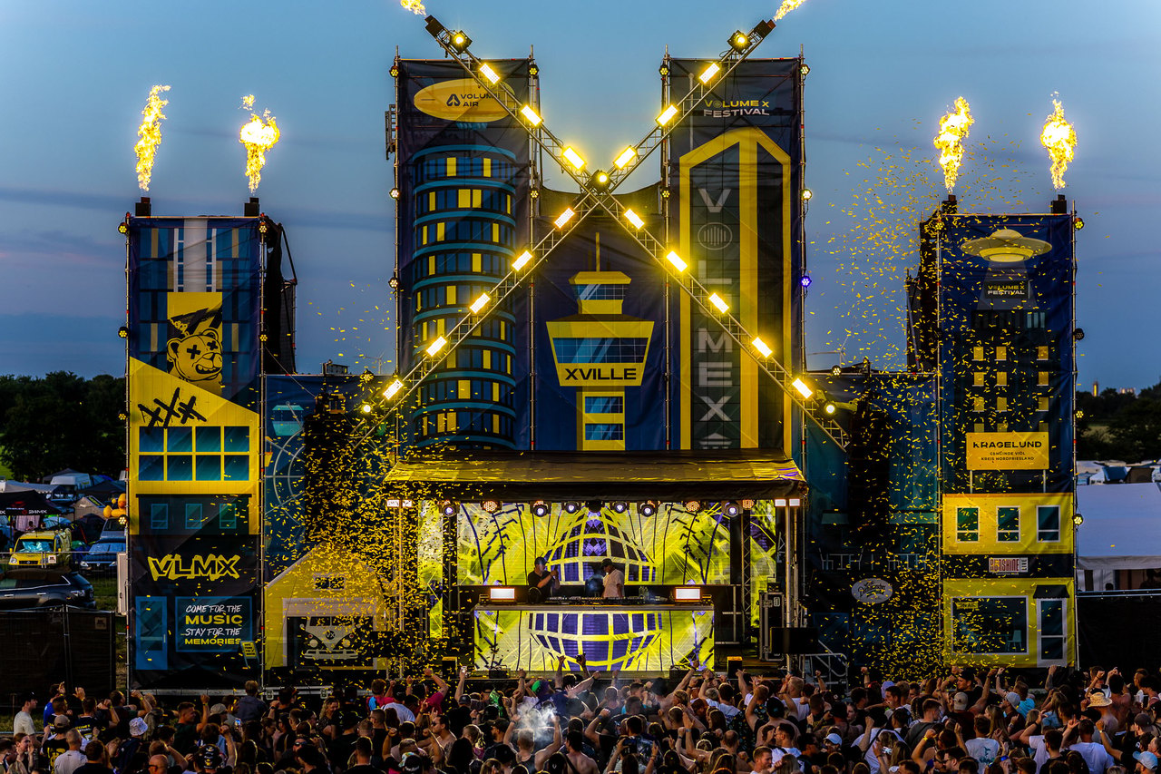 DJ-Bühne im Stadtbild-Design mit Feuereffekten und Konfetti, davor eine Menschenmenge.